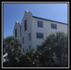 White building with blue windows and palm trees against a clear blue sky.