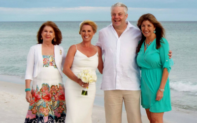 Four adults posing on a beach, one holding a bouquet, with ocean in the background.