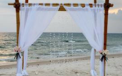 White beach wedding arch with curtains and flowers by the ocean at sunset