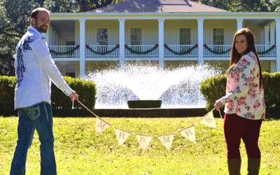 Couple holding a “BABY” banner in front of a fountain and white house with garlands