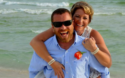 Smiling couple embracing on a beach, with waves in the background and the man wearing sunglasses