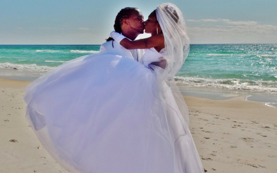 Bride in a white gown and veil kisses another person on a sandy beach by the ocean.