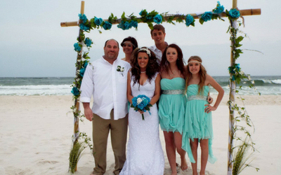 Wedding group posing on a beach under a decorated arch, bride in white with attendants in mint dresses