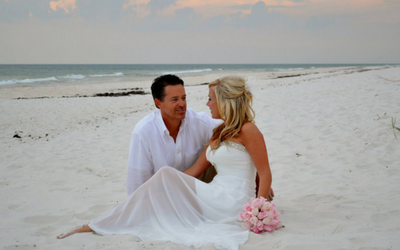 Bride and groom sitting on a white-sand beach at sunset, facing each other with pink bouquet nearby