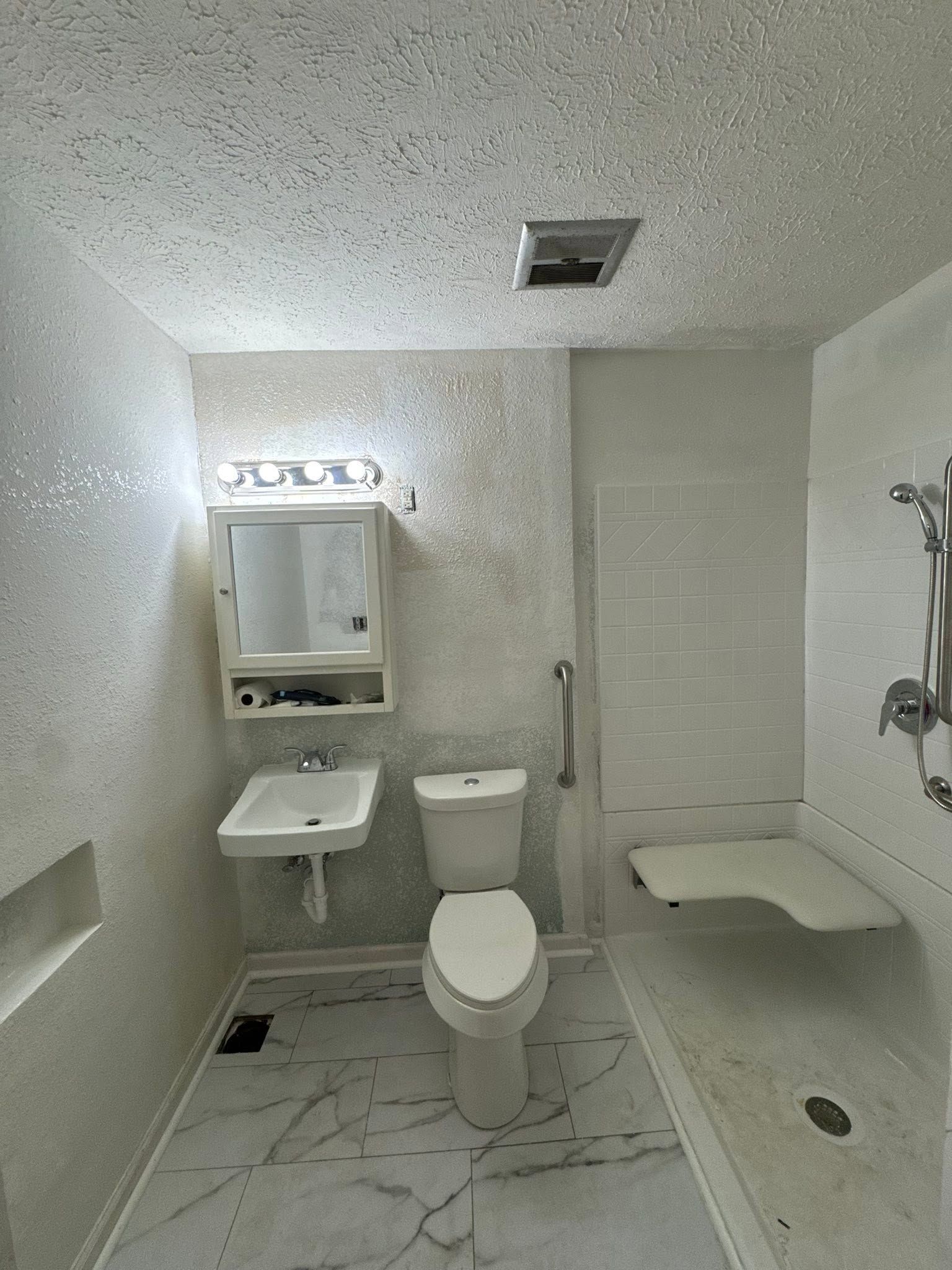 Small white accessible bathroom with sink, toilet, shower seat, and grab bars