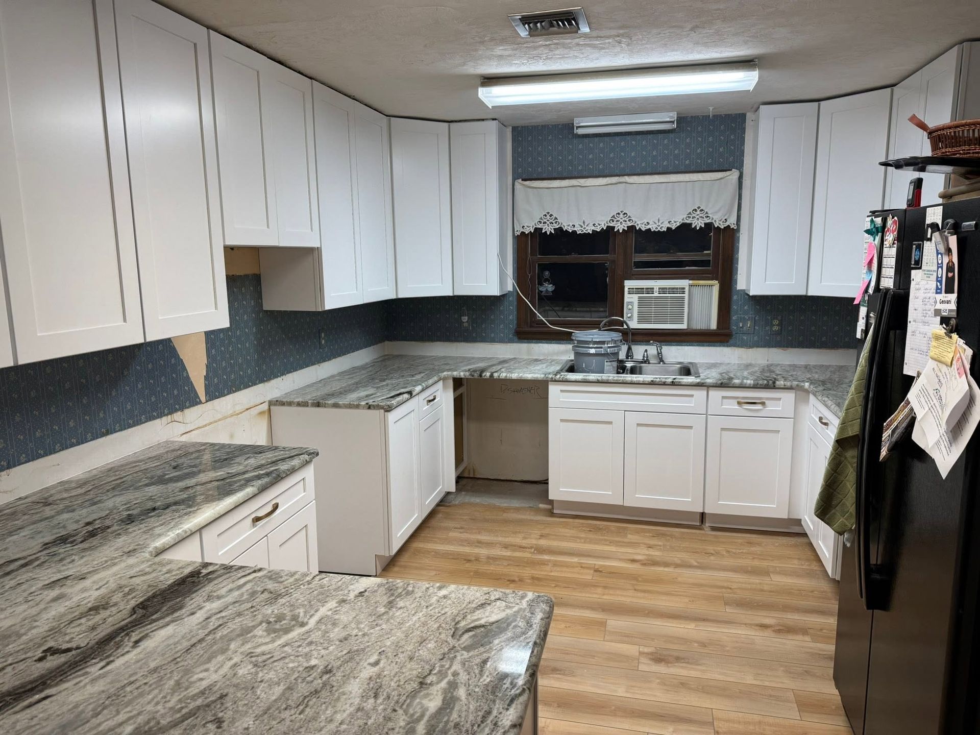 Small kitchen with white cabinets, black countertops, wood floor, and a window air conditioner above the sink.