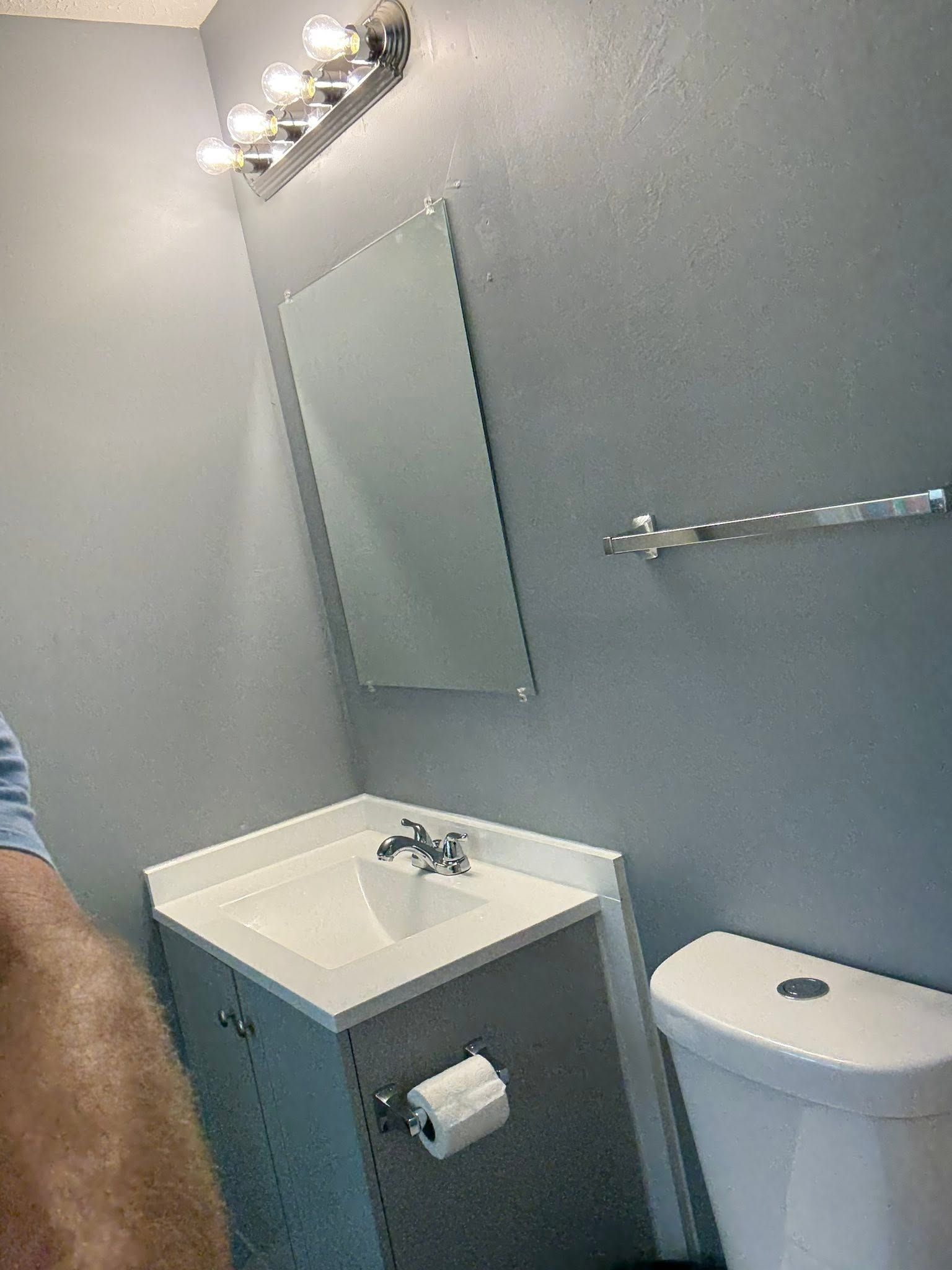Small gray bathroom with sink, mirror, toilet, towel bar, and a person’s arm at left.