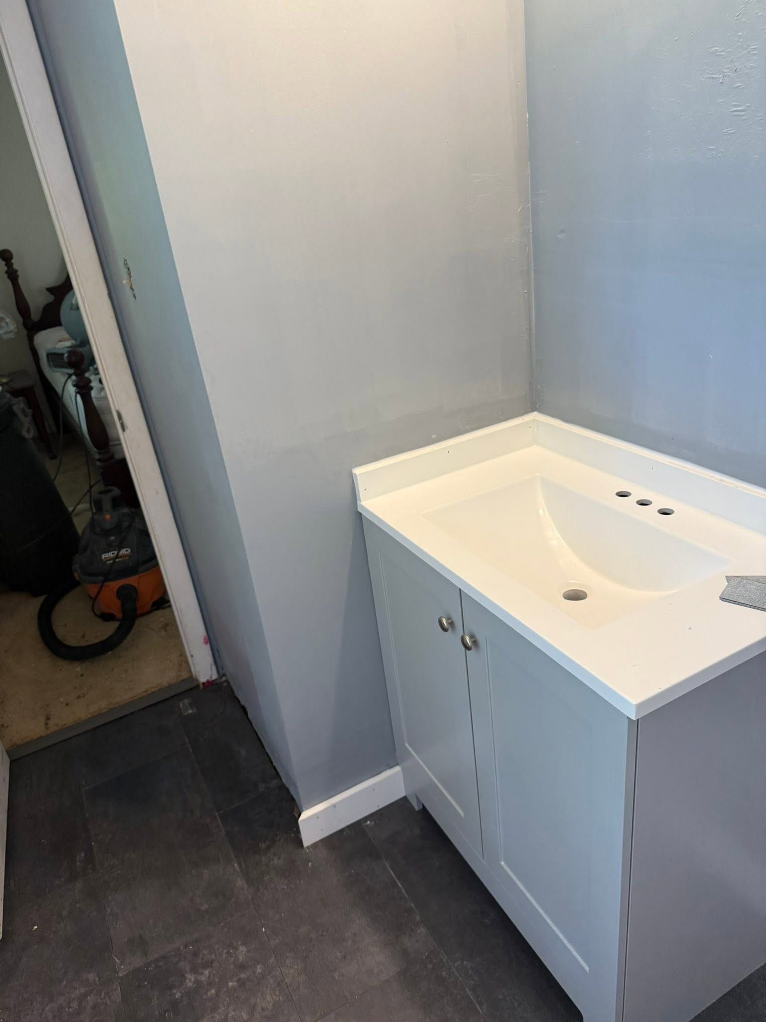 Small white bathroom vanity with sink in a narrow room with gray walls and dark tile floor