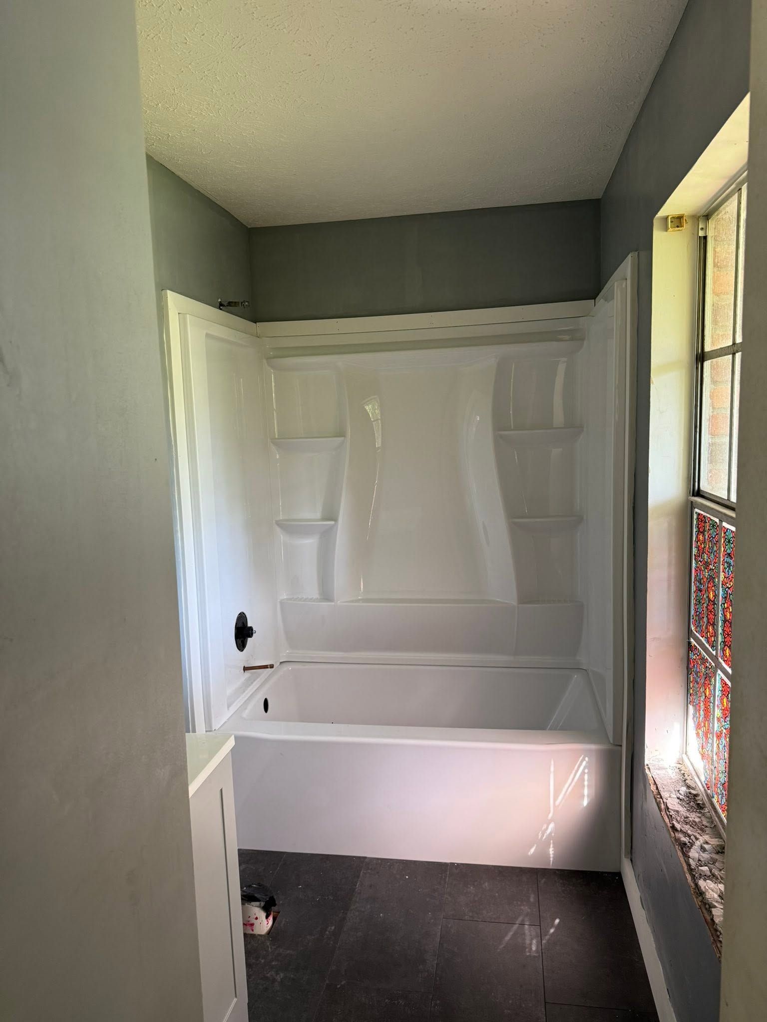 Empty bathroom with a white bathtub/shower surround, gray walls, and a window on the right.