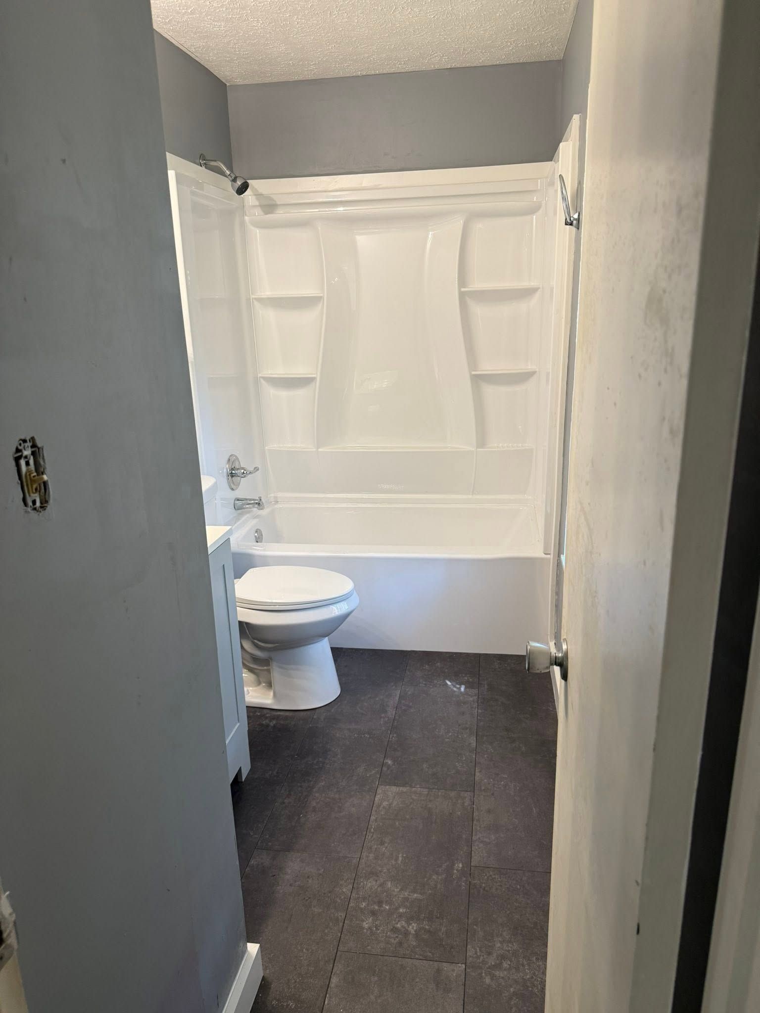 Narrow bathroom hallway with a toilet and bathtub at the end, gray walls and dark floor tiles.