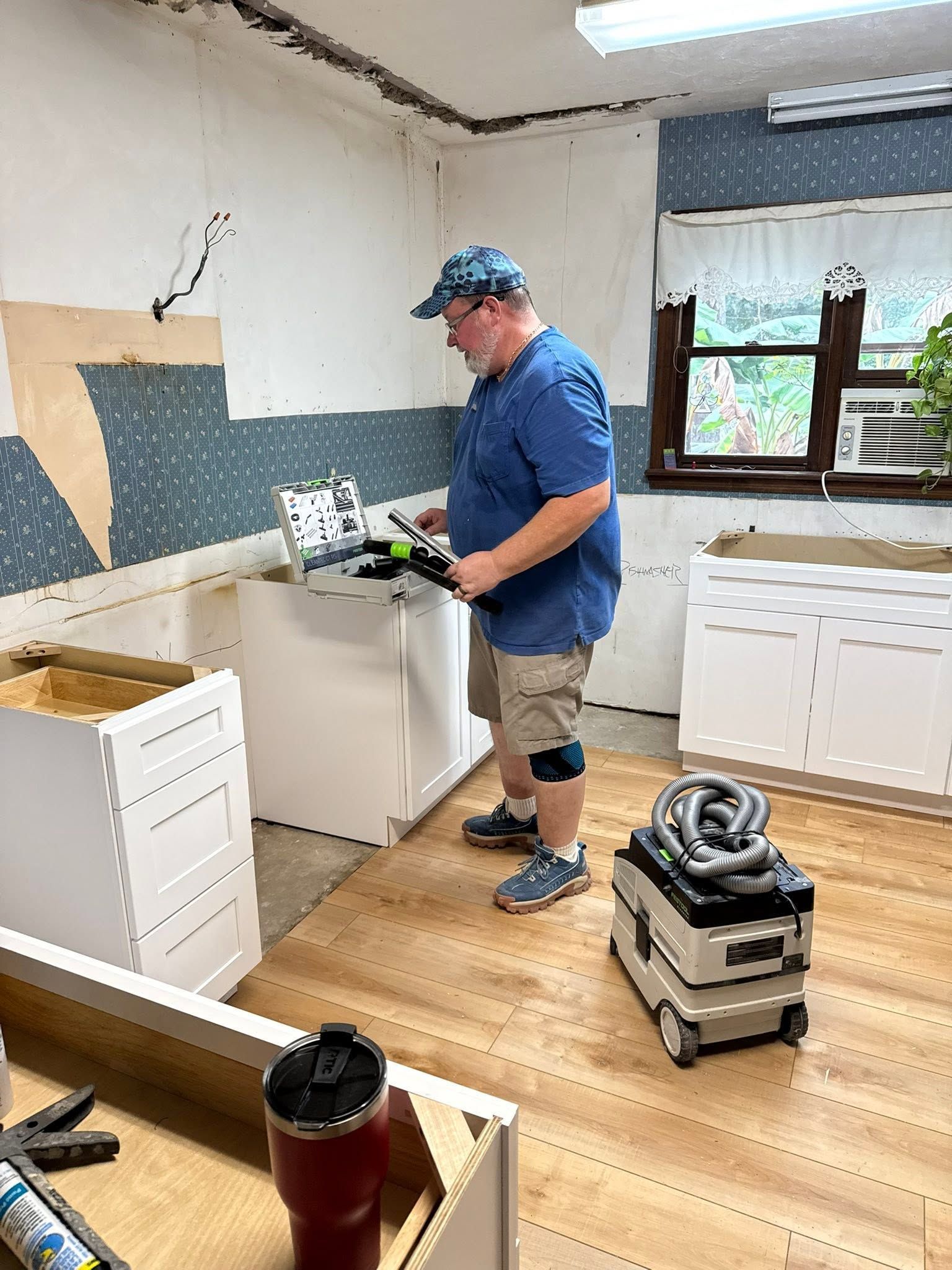 Person working on kitchen renovation beside cabinets and tools in a room with exposed walls and wooden floor