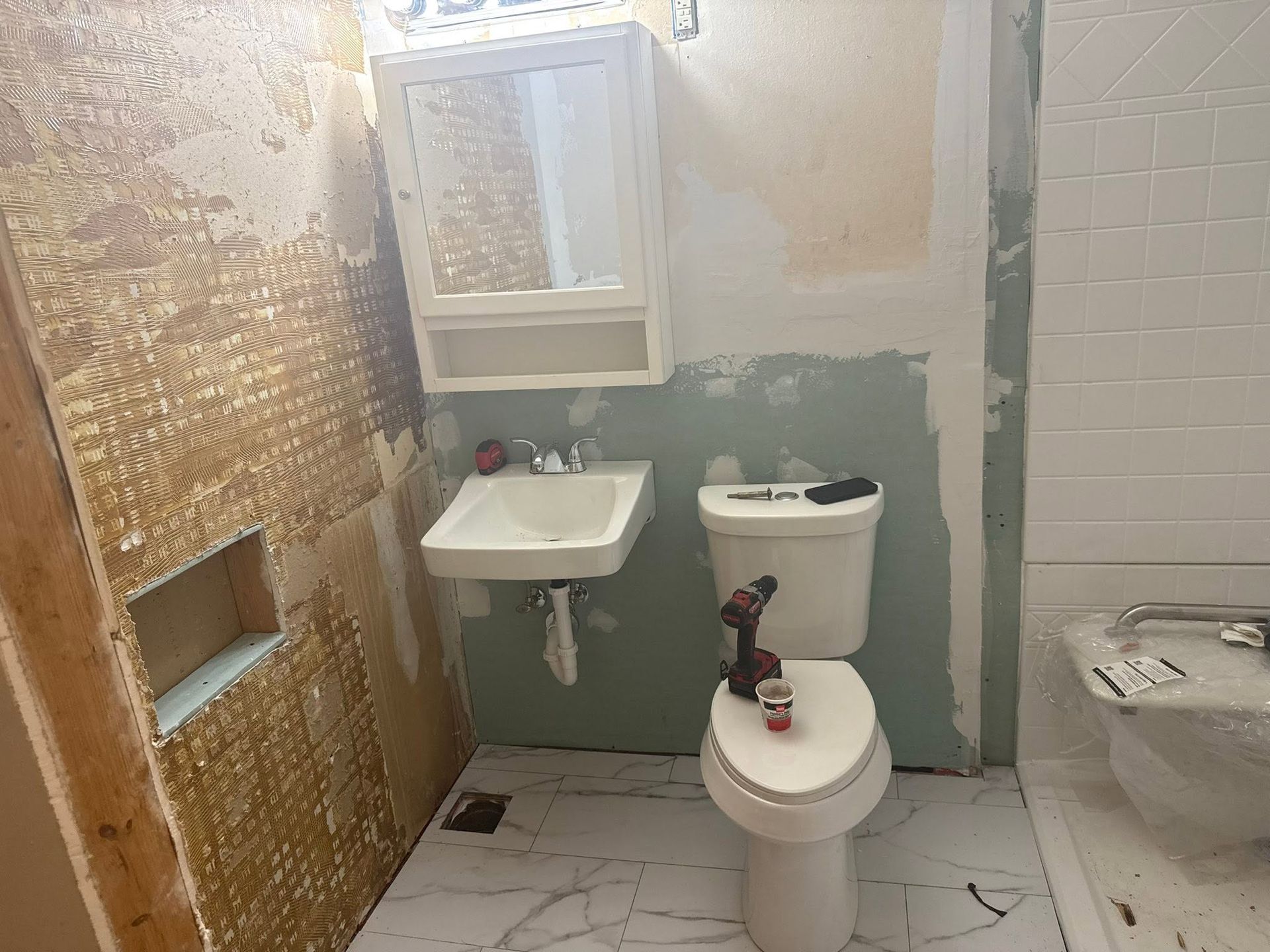 Small bathroom with toilet, sink, mirror, and a partially installed tub in a rough unfinished room.