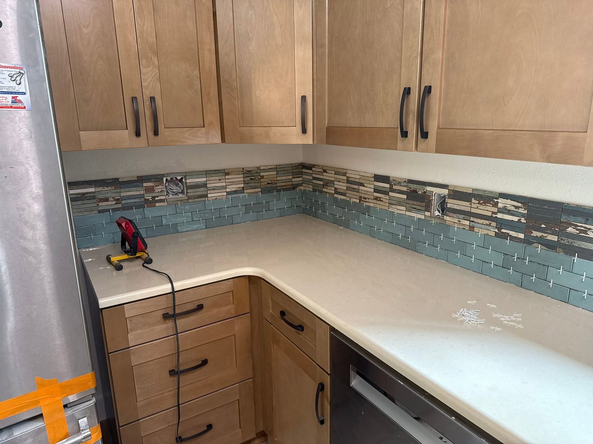 Small kitchen with light wood cabinets, white countertop, mosaic backsplash, and a cordless drill on the counter