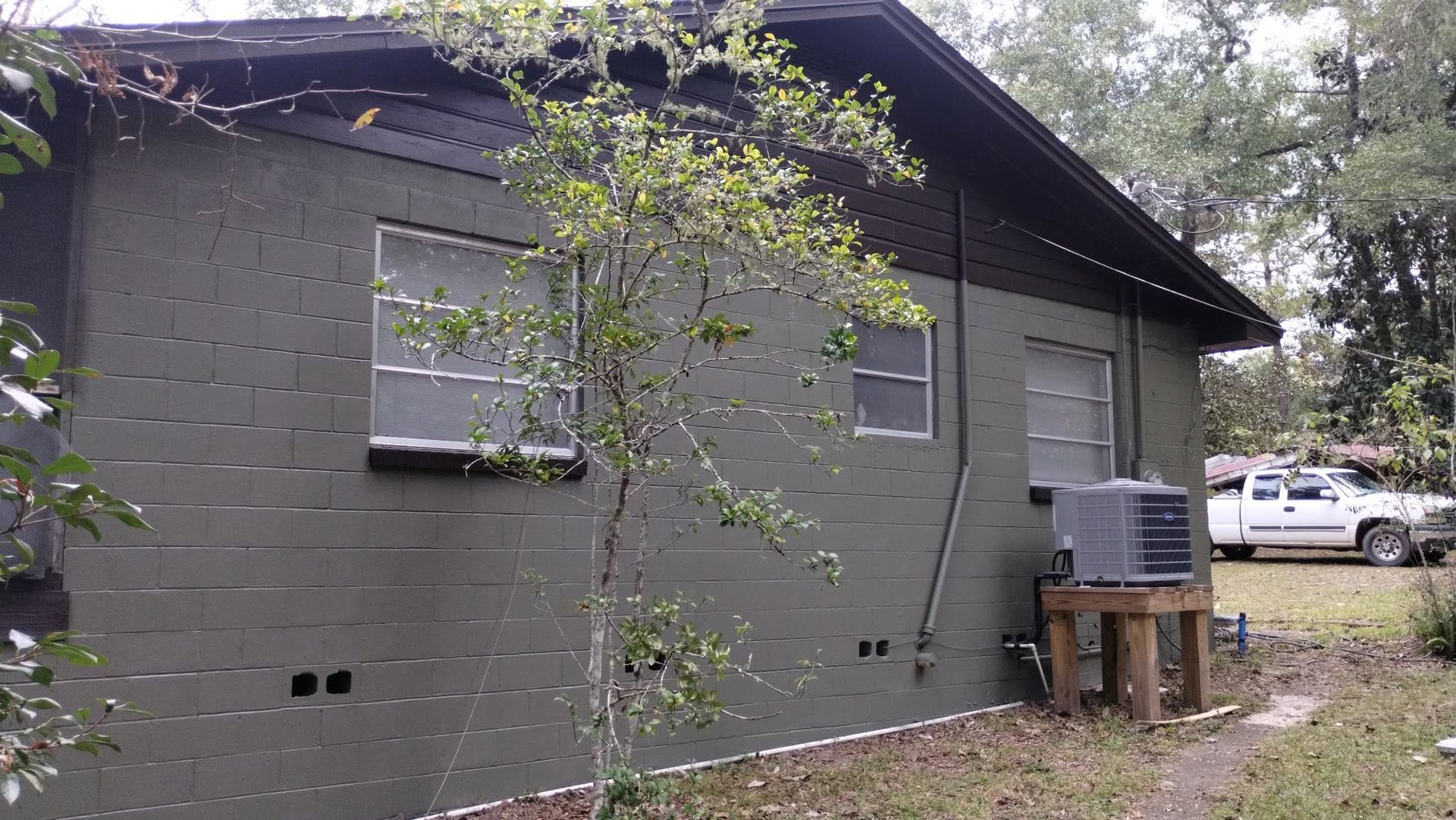 Gray house exterior with windows, a tree in front, and an HVAC unit on a wooden platform.