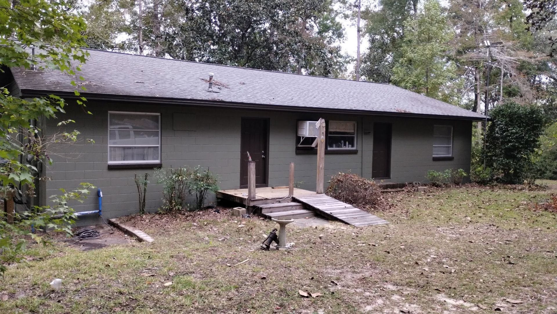 Small gray single-story house with a front porch and ramp, surrounded by trees and fallen leaves.