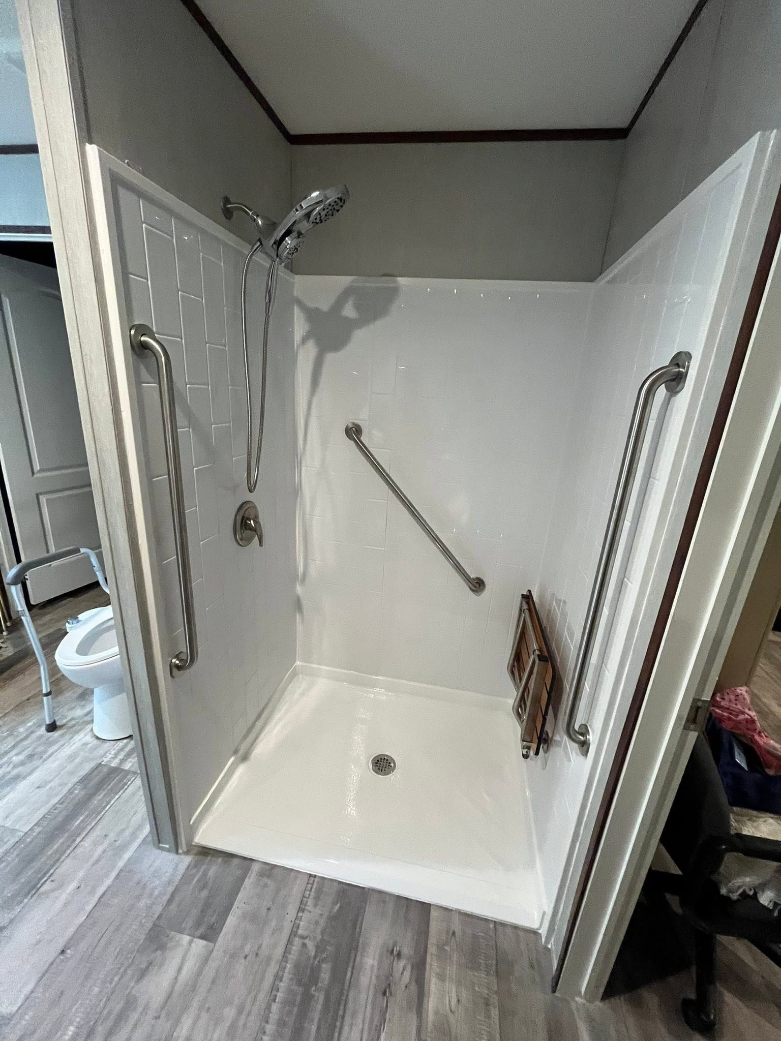 Accessible shower stall with white walls, grab bars, fold-down seat, and handheld showerhead in a bathroom.