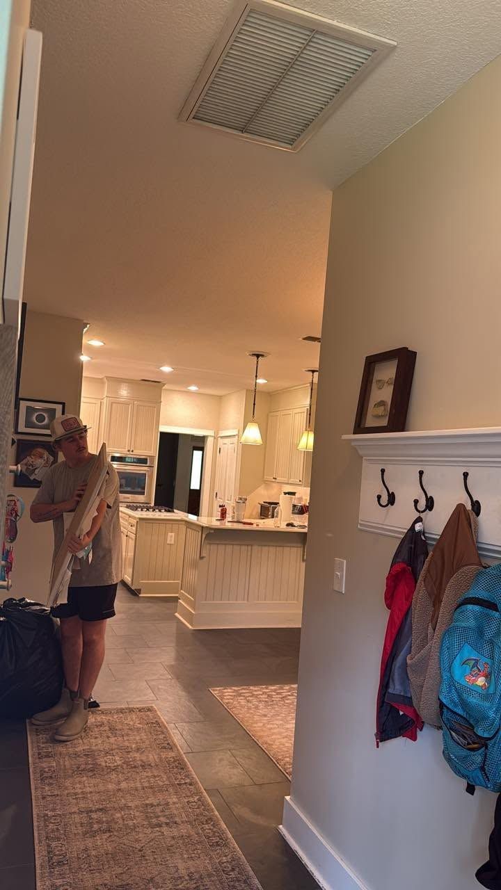 Hallway leading to a bright kitchen; a person stands by the wall near a coat rack and rug.