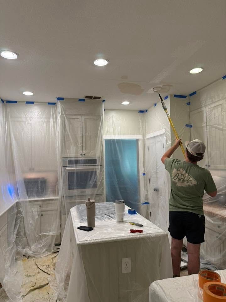 Person painting a room ceiling while walls and windows are covered in plastic sheeting.