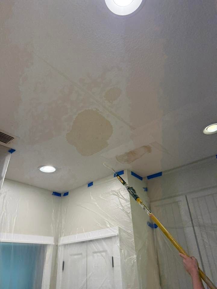 Person painting a ceiling with a roller, covering water stains in a white room