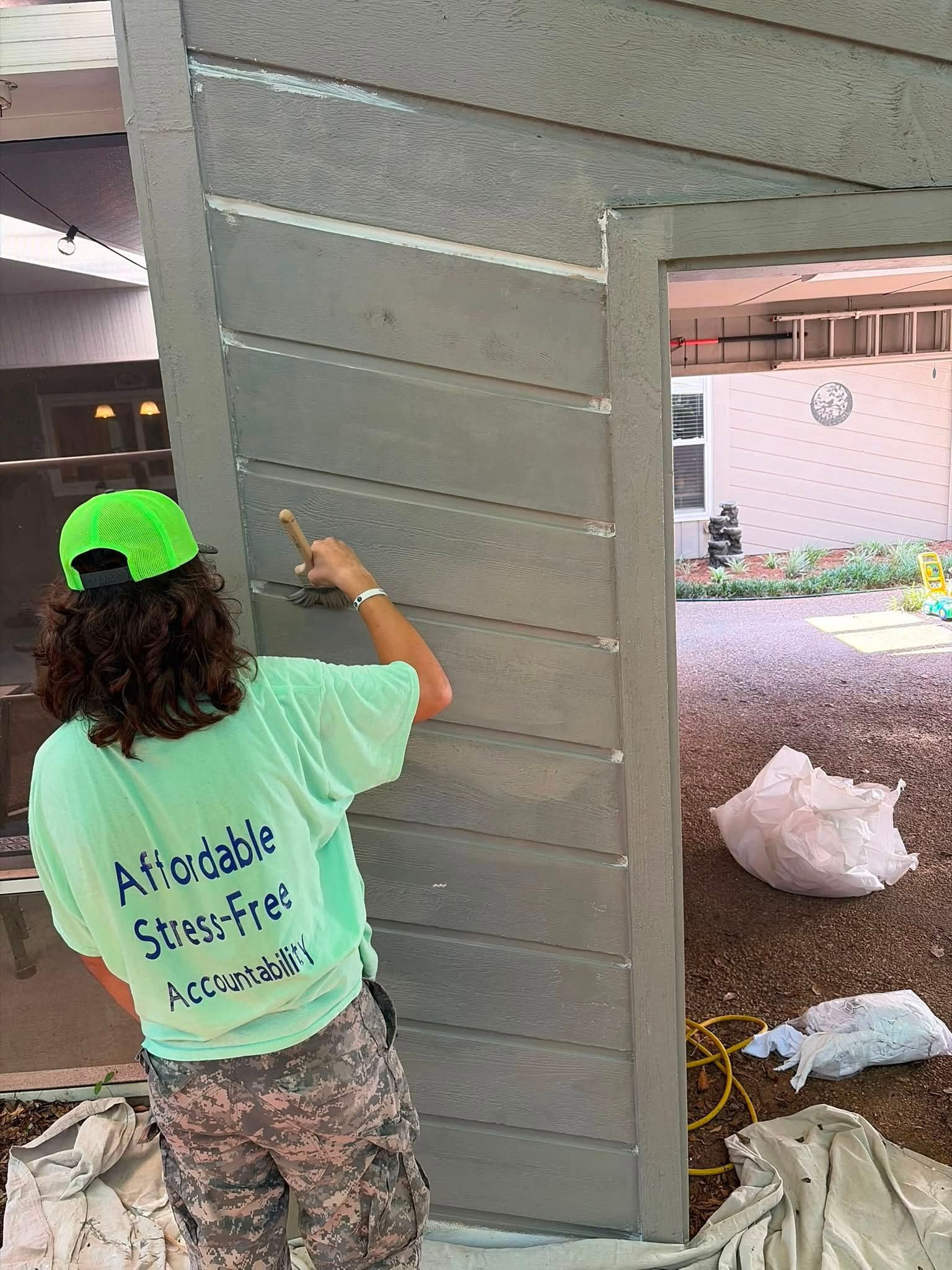 Person painting a gray wall panel outdoors near a building entrance, wearing a neon green cap and camouflage pants