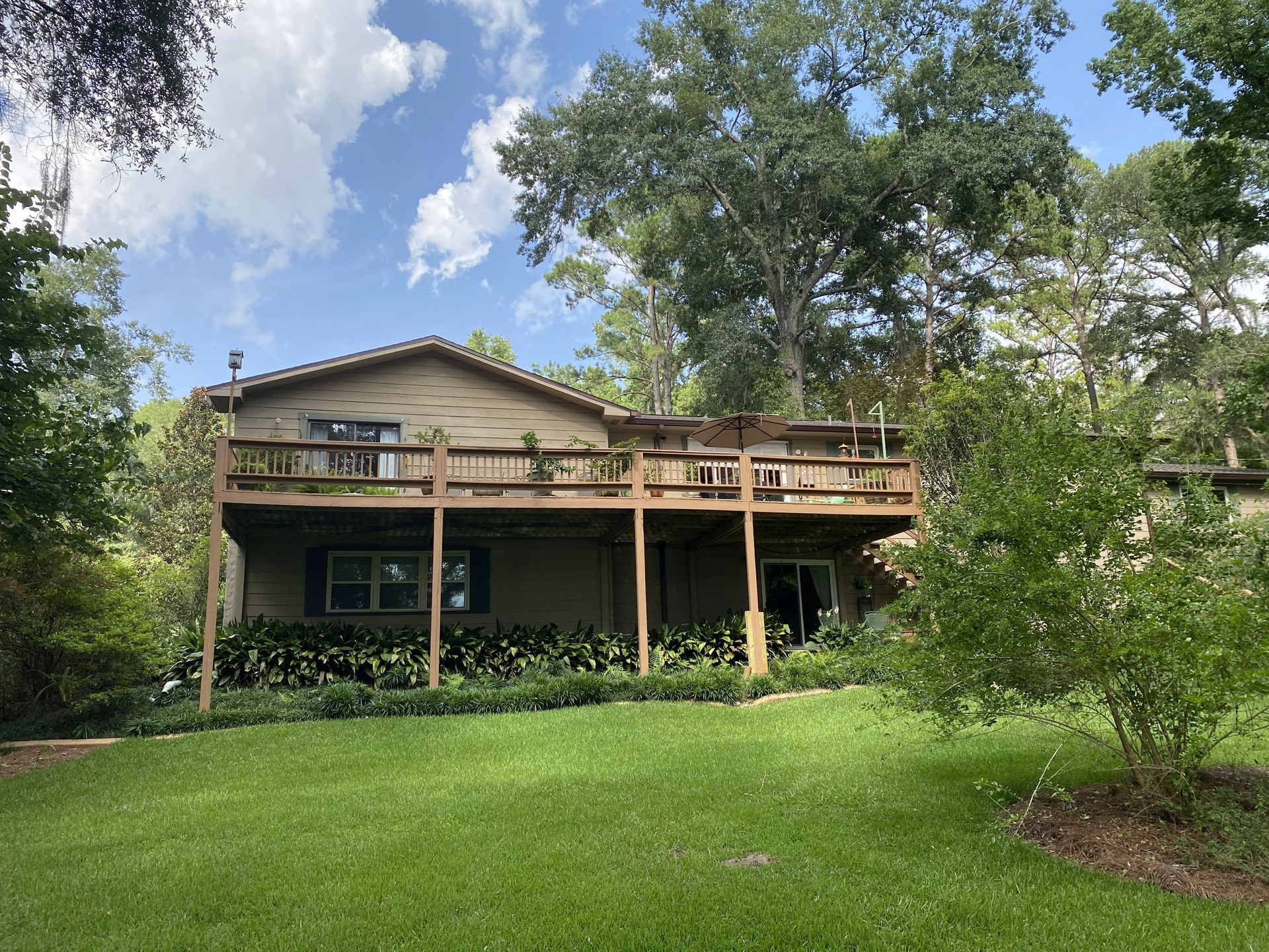 Two-story house with a wooden deck, surrounded by trees and a green lawn under a partly cloudy sky.