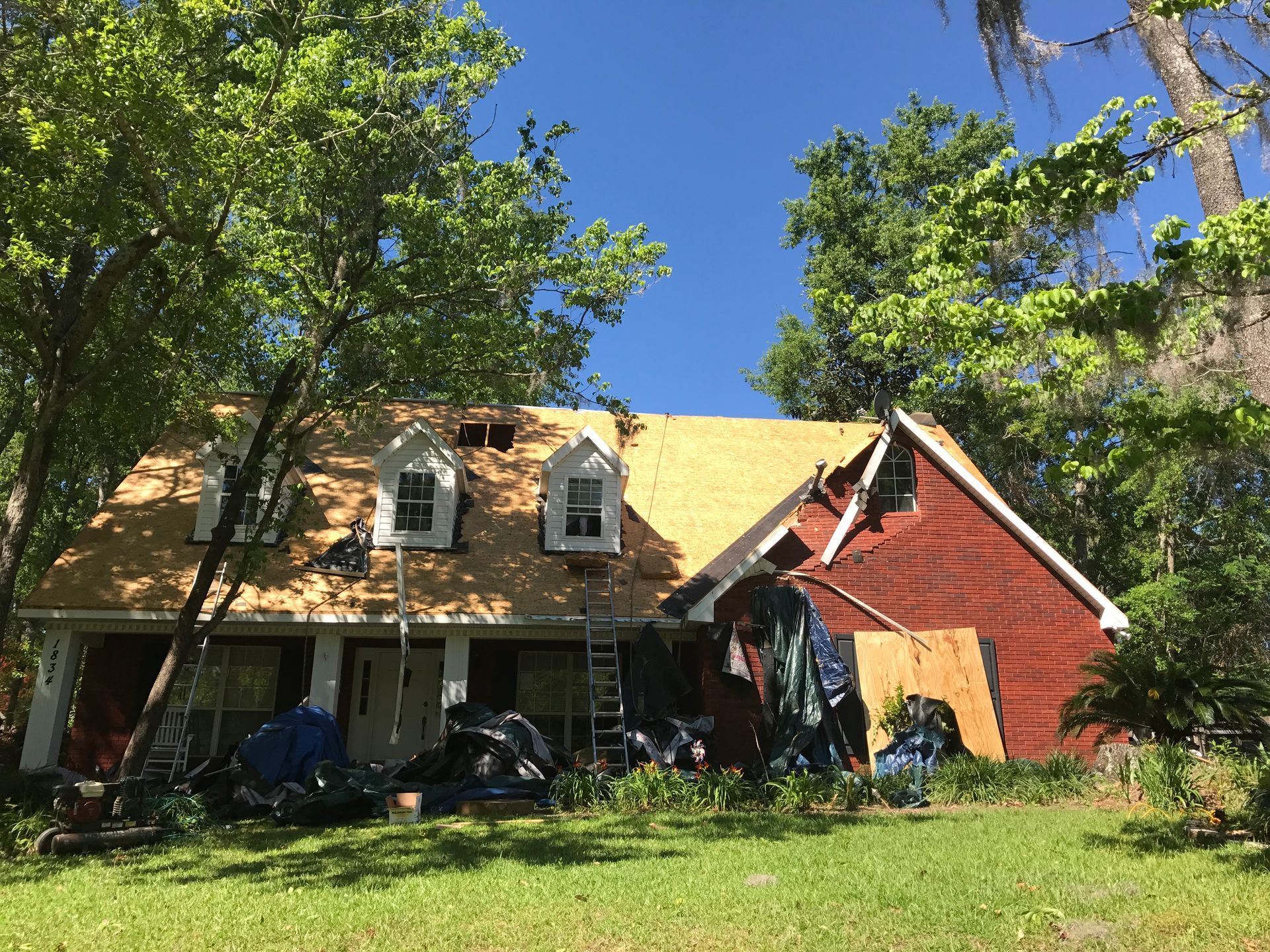 Red house with tan roof amid trees and blue sky, front porch partially hidden by lawn and foliage
