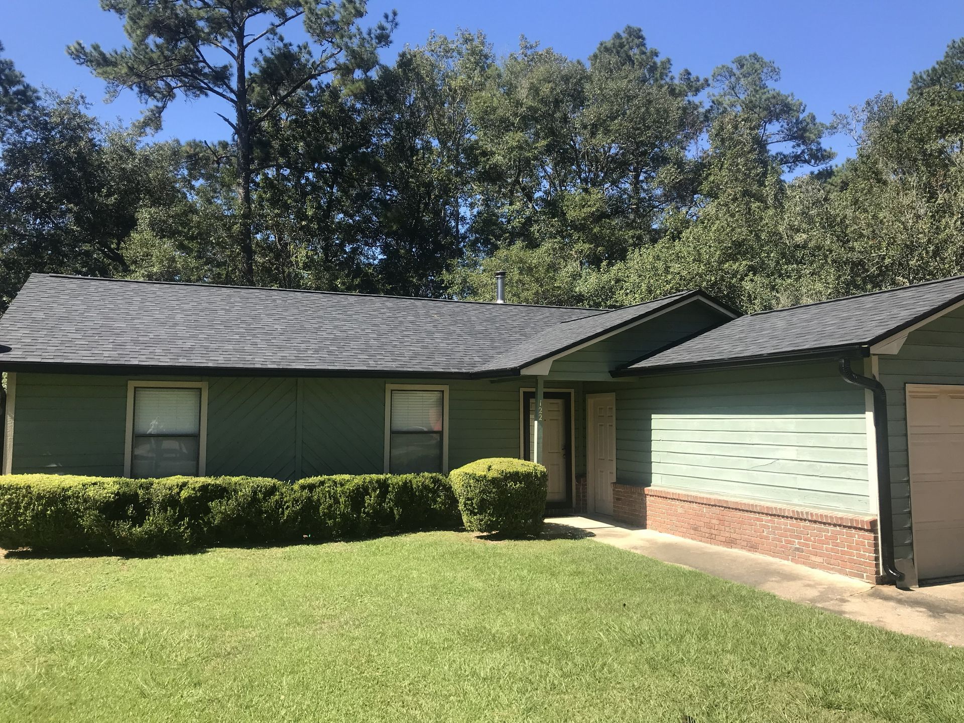 Single-story green house with dark roof, brick trim, and a front lawn under trees.