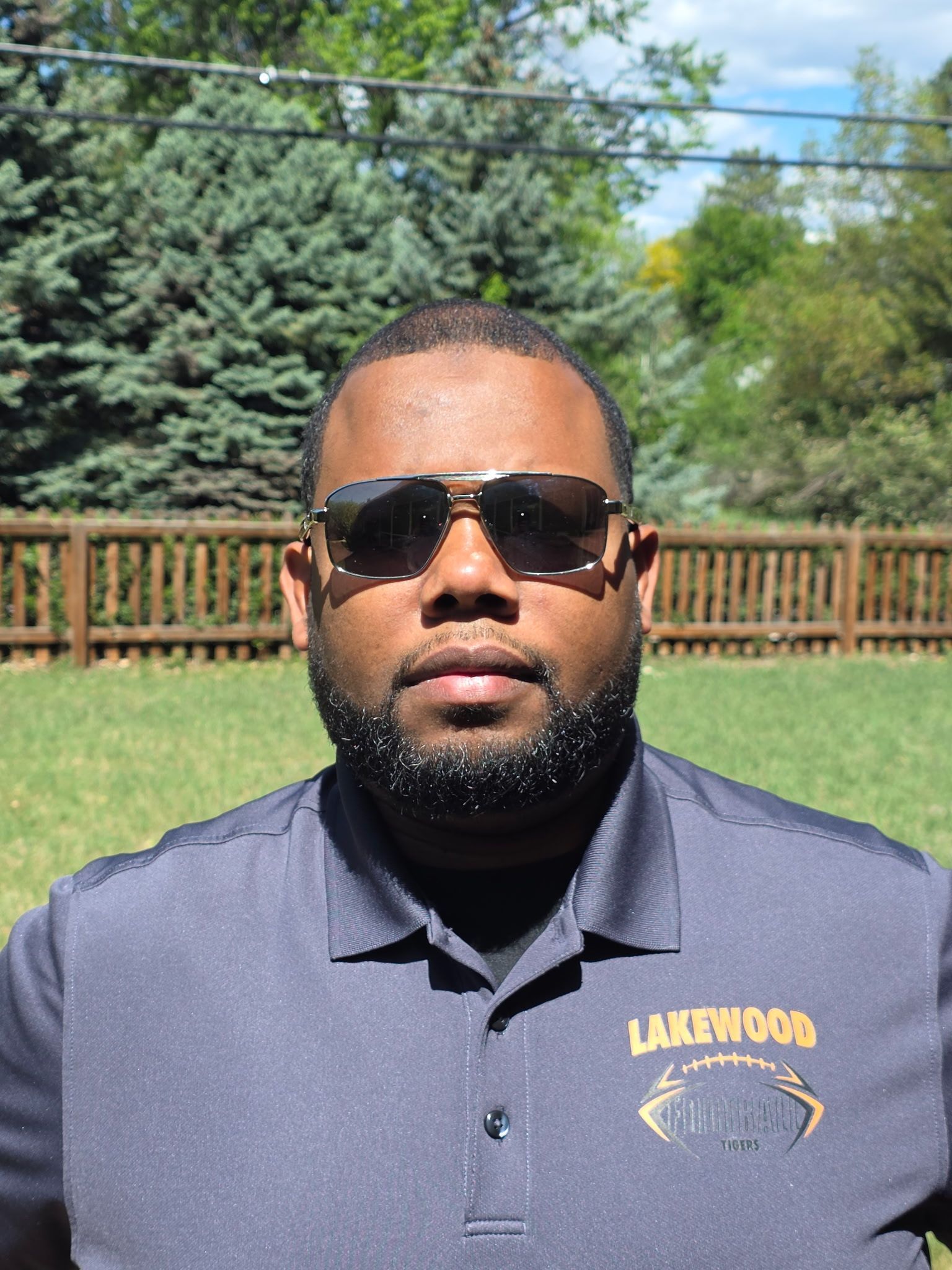 Man wearing sunglasses and a navy polo stands outdoors in front of a wooden fence and trees.