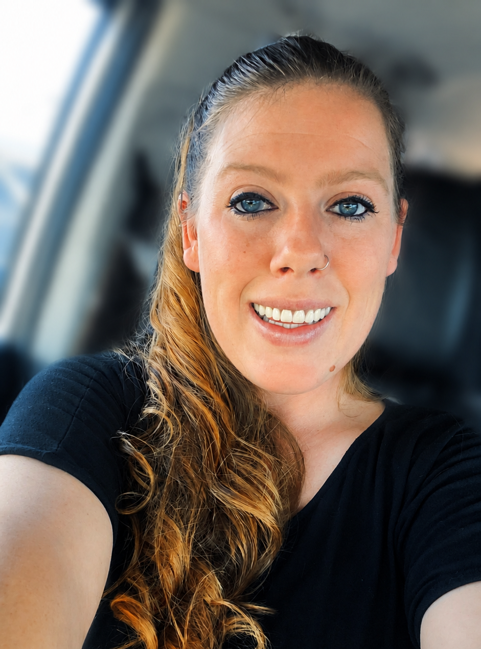 Woman smiling in a car, wearing a black shirt with long hair pulled back.
