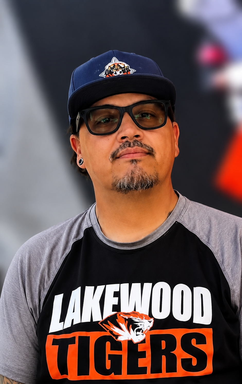 Man in a black and gray LAKEWOOD TIGERS shirt, navy cap, and sunglasses, standing at an event