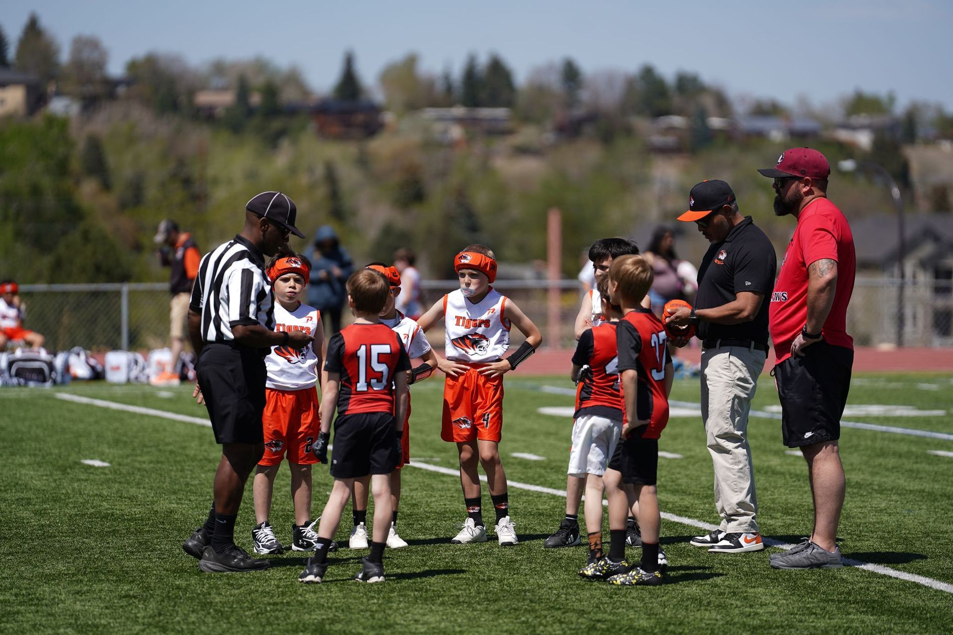 Youth lacrosse huddle on a sunny field with players in orange, black, and white uniforms and coaches nearby