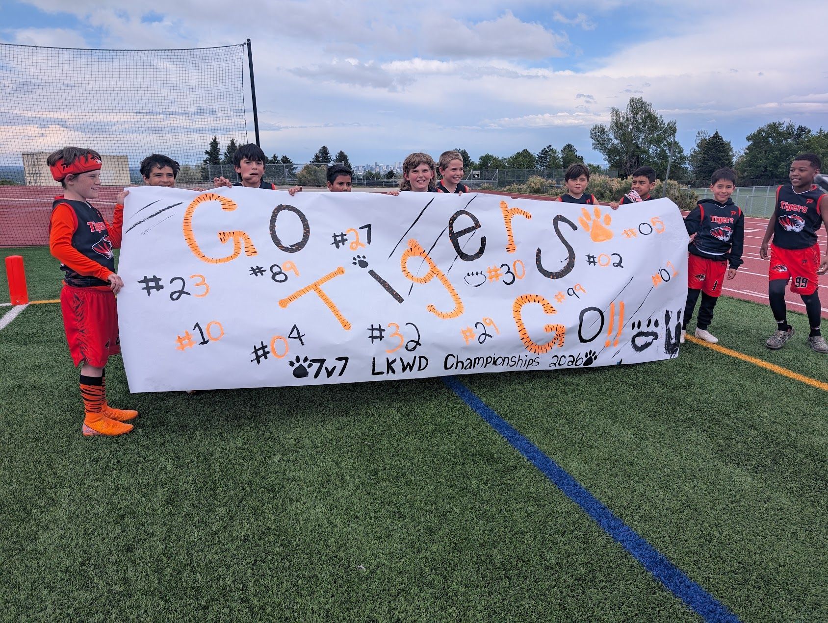 Youth soccer team holding a white banner on a turf field, with goal net and cloudy sky behind them.