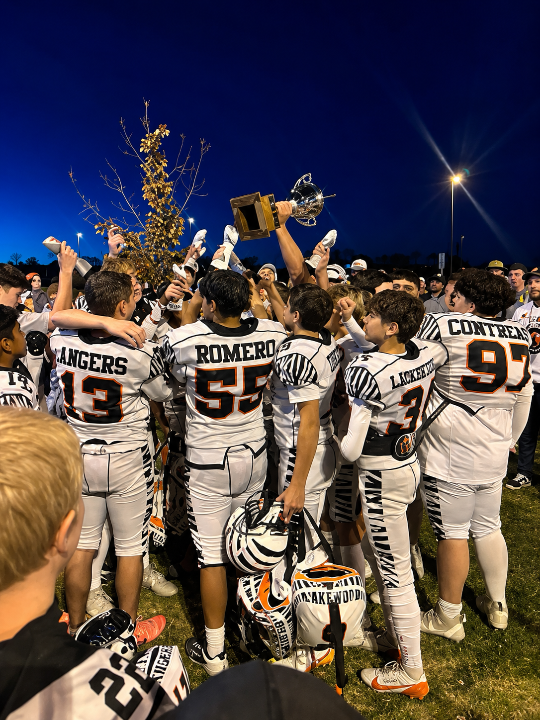 High school football team in white uniforms cheering and lifting a trophy at night on a field