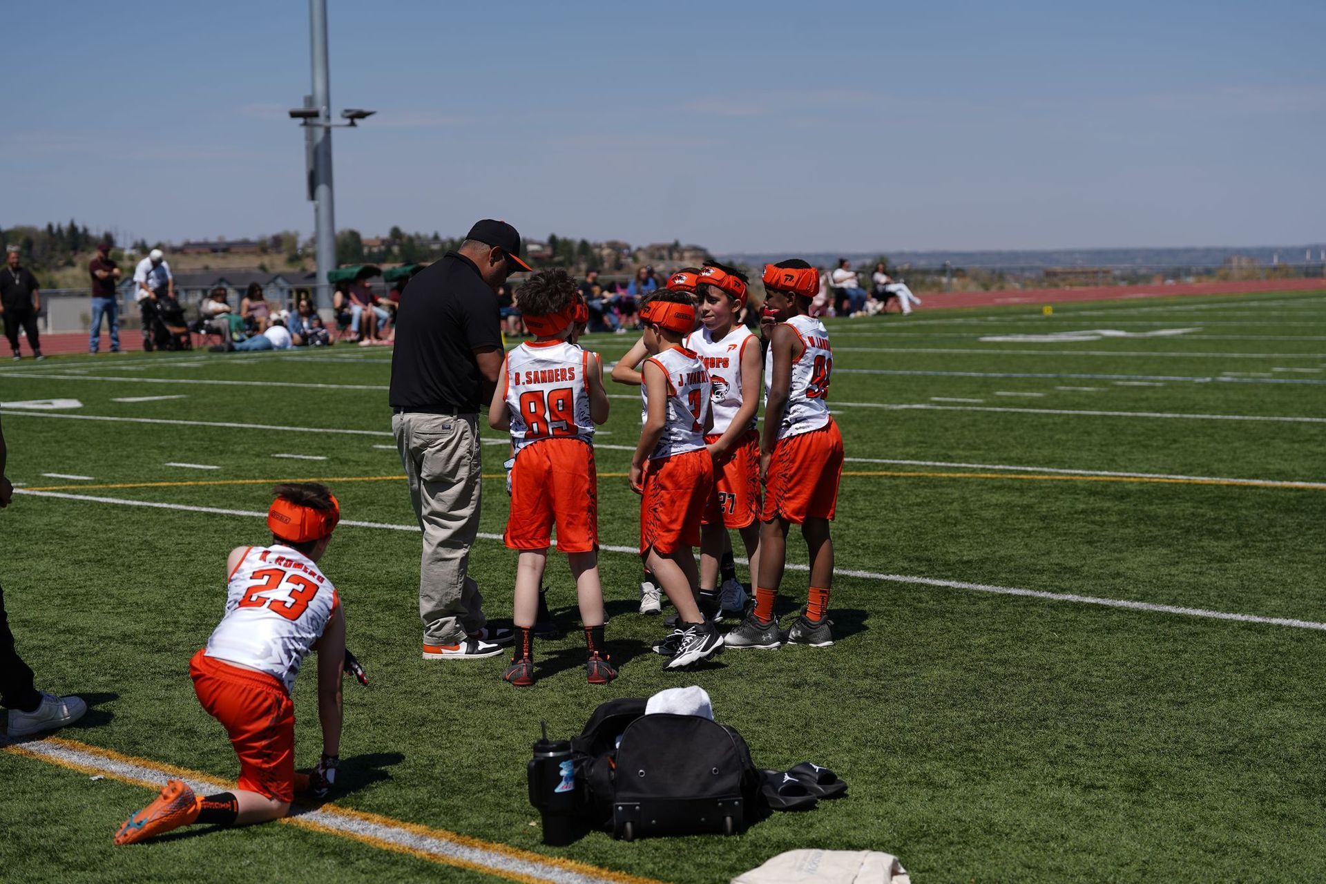 Youth football players in orange and white huddle on a field during a game; one kneels nearby.