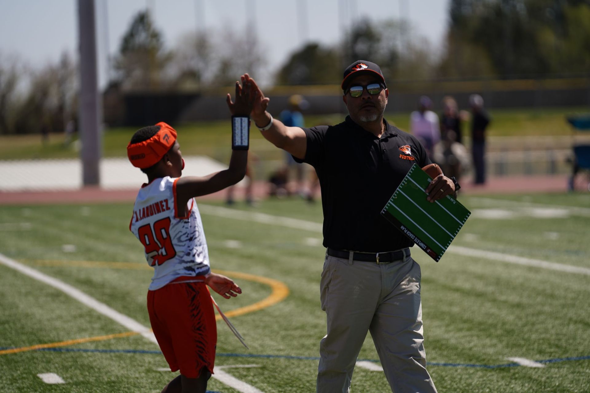 Two football players high-five on a field, one holding a green play board.