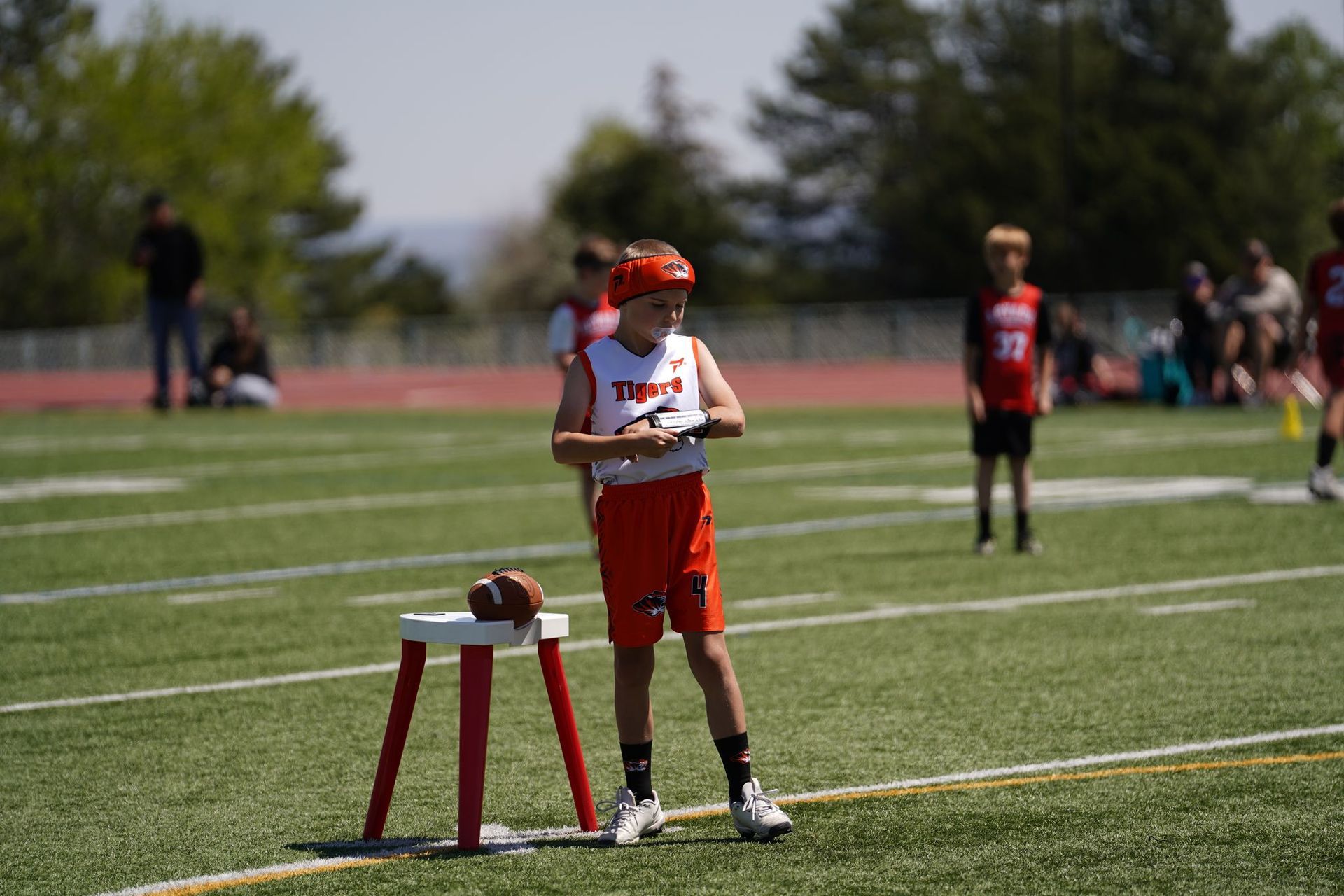 Boy in red baseball uniform standing beside a chair on a sports field, holding a paper in bright sunlight.