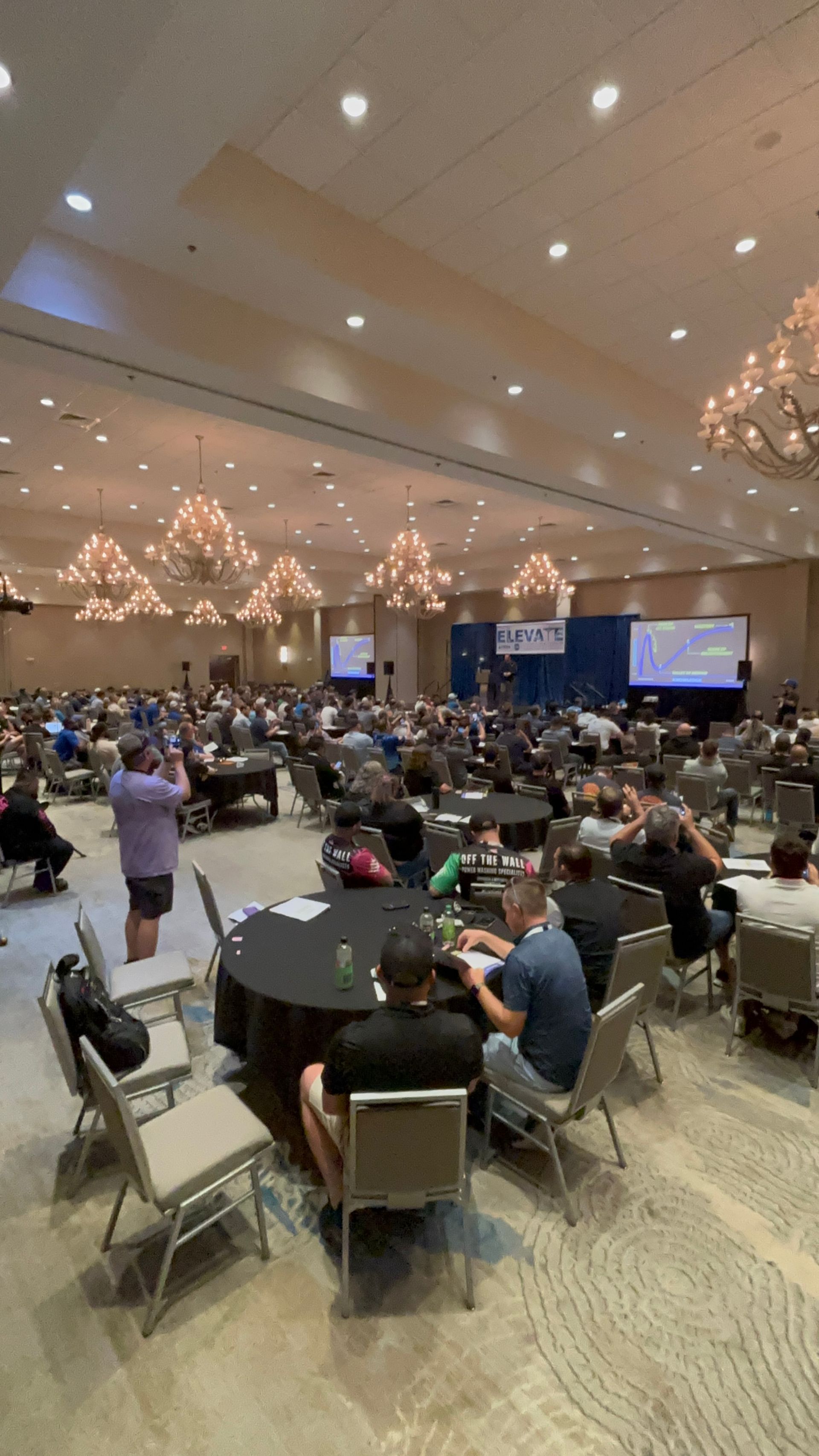 Large banquet hall filled with round tables and seated attendees facing a stage with screens and chandeliers