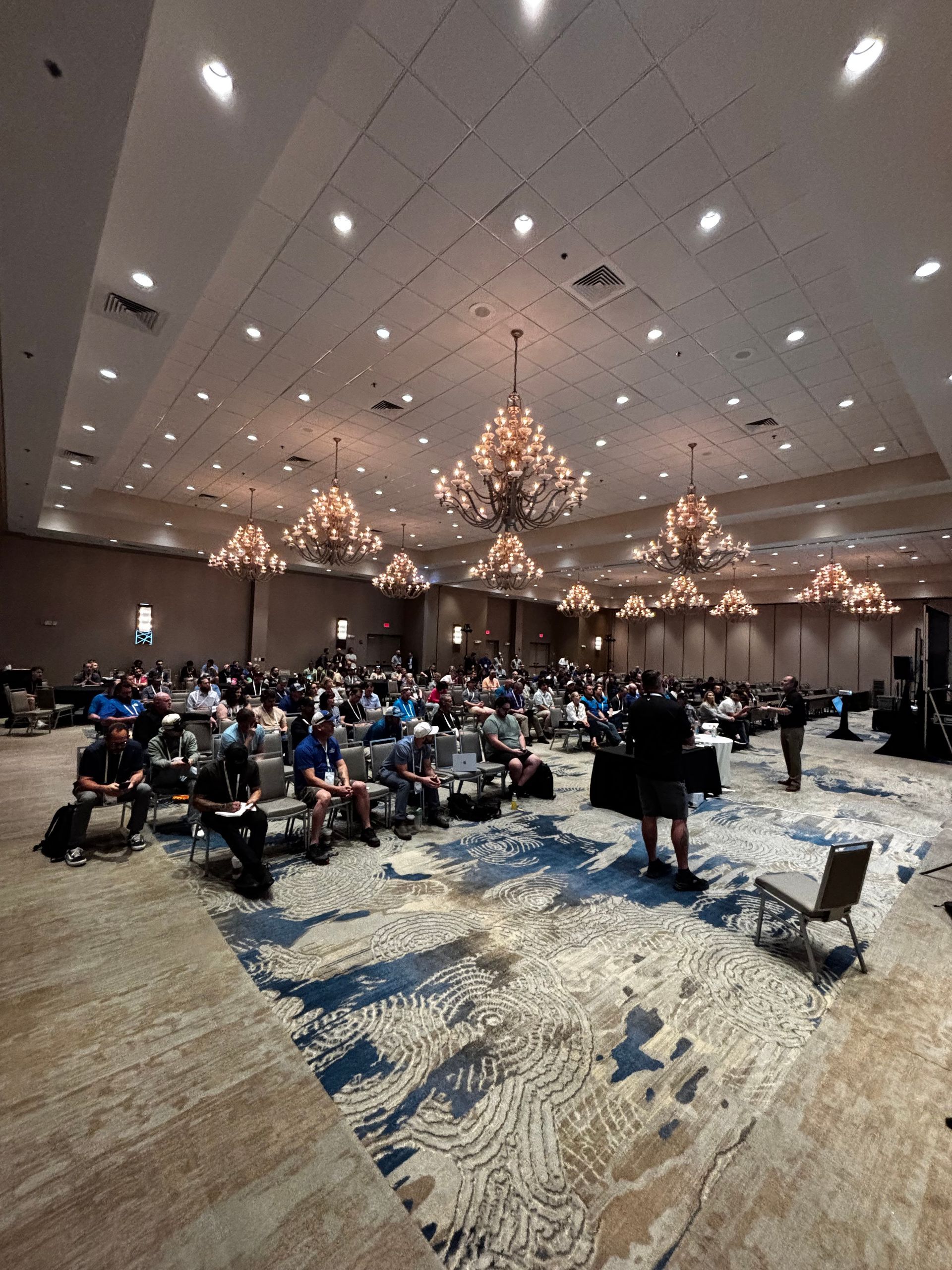 Large banquet hall with chandeliers and many people seated on the floor, possibly for an event or ceremony.