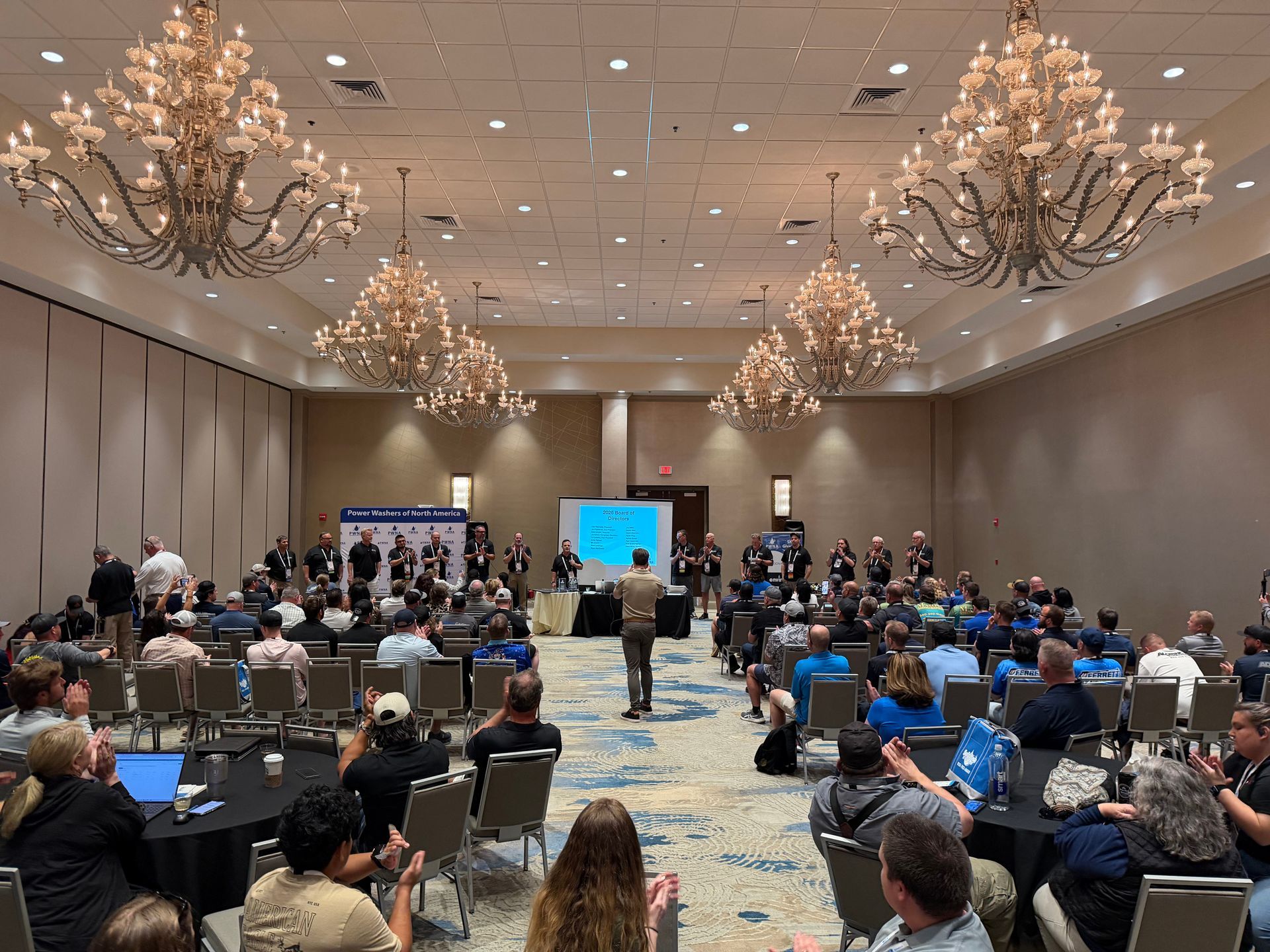 Conference audience facing a presentation screen in a large ballroom with chandeliers and round tables.
