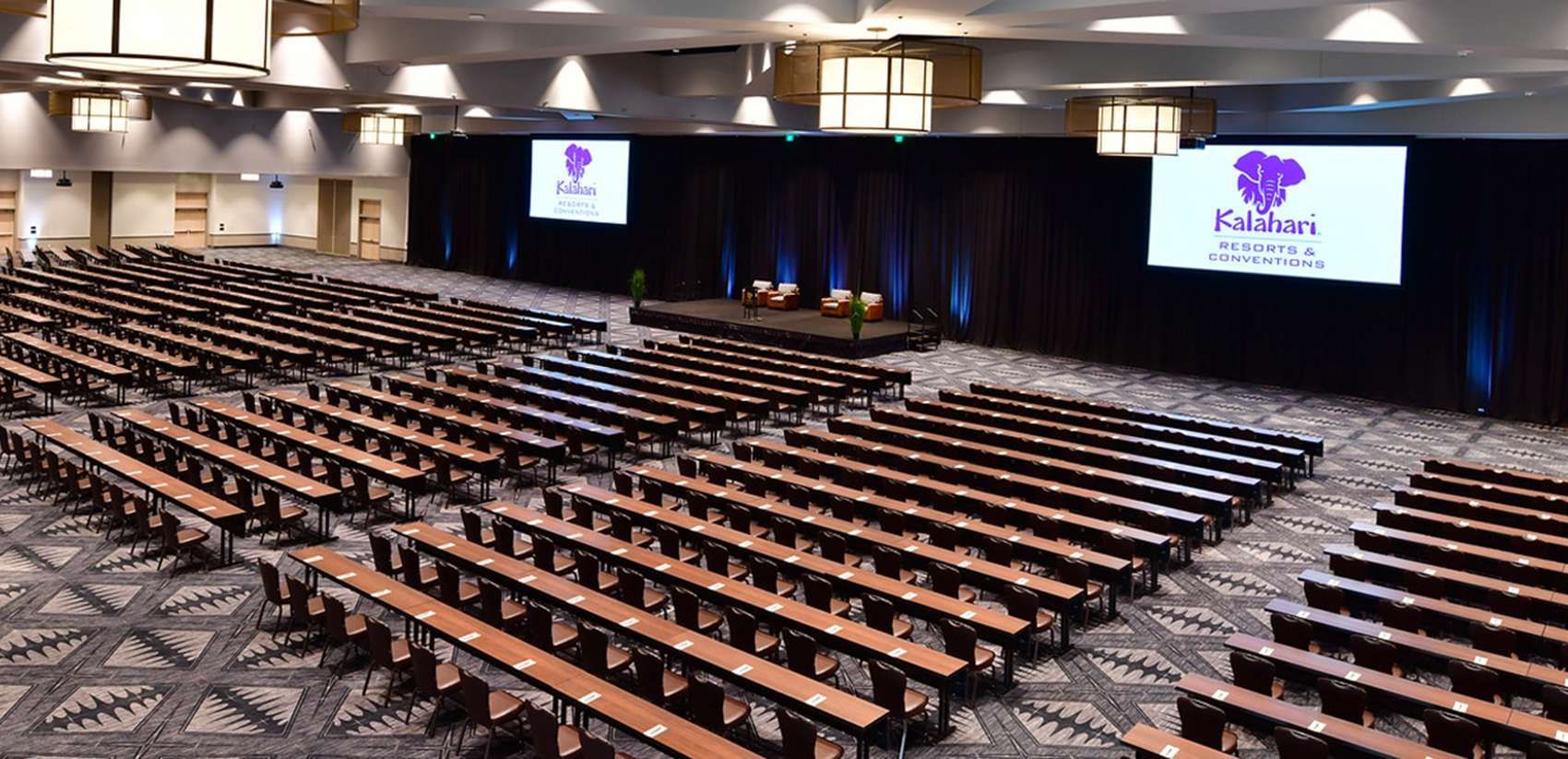 Large conference auditorium with rows of long tables facing a stage and screens