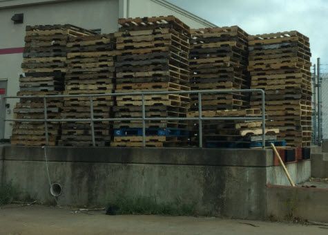 Stacked wooden pallets piled high behind a concrete wall in an industrial area.