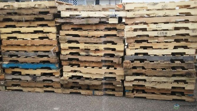 Stacked wooden pallets in a warehouse yard, with some blue-painted pallets among mostly beige and brown ones