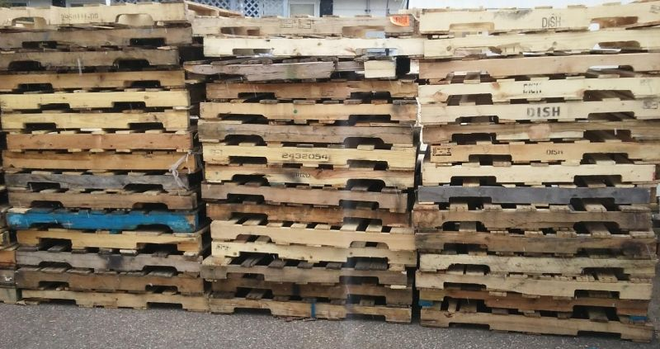 Stacked wooden pallets against a wall, with some blue-painted ones mixed in.