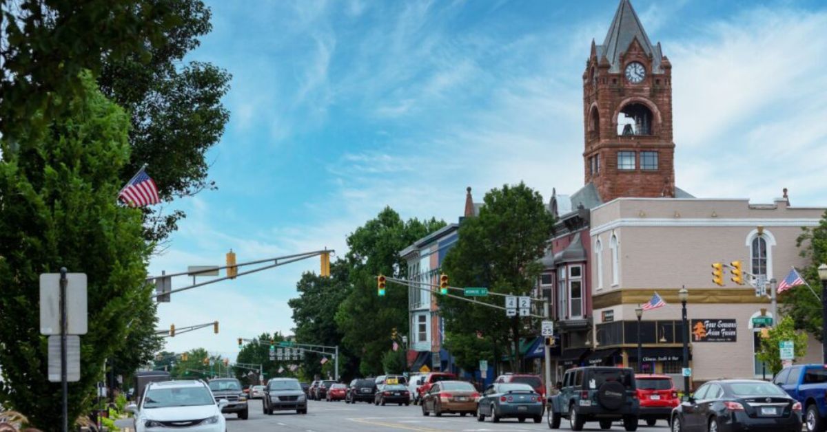Downtown street with cars, trees, storefronts, and a red brick clock tower under a blue sky