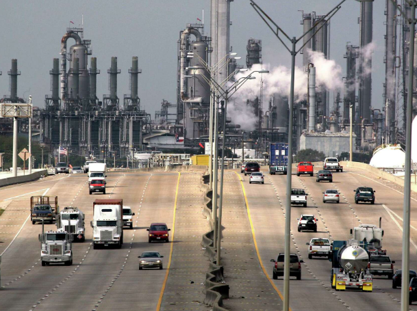 Heavy traffic on a highway beside a large industrial refinery with smokestacks and steam.