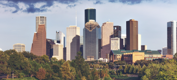 City skyline with modern skyscrapers above green trees under a partly cloudy sky