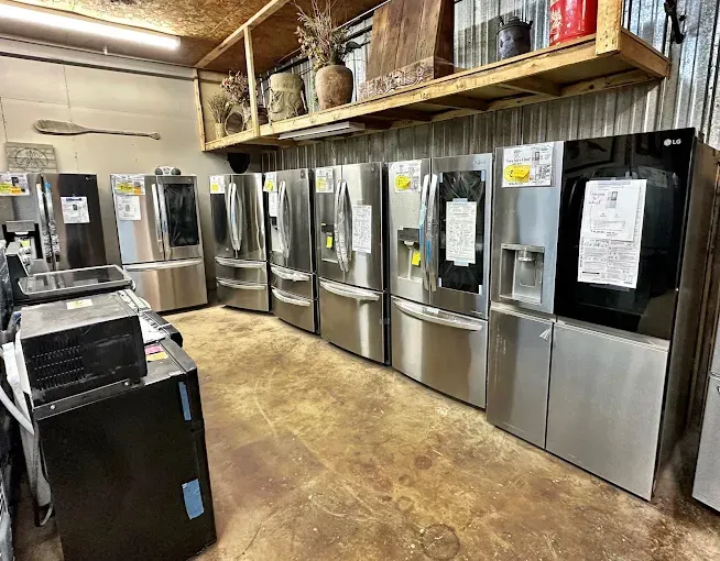 Row of stainless steel refrigerators in a warehouse-style appliance showroom with concrete floor and wooden shelves above