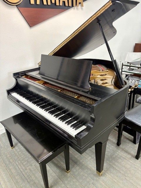 Black grand piano with lid open and bench in a showroom with other pianos in the background