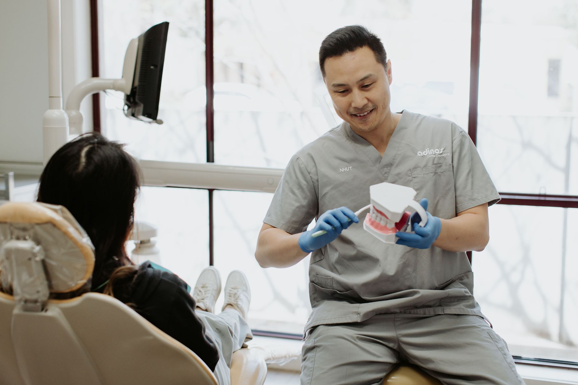 A dentist is holding a model of a tooth while talking to a patient in a dental chair.