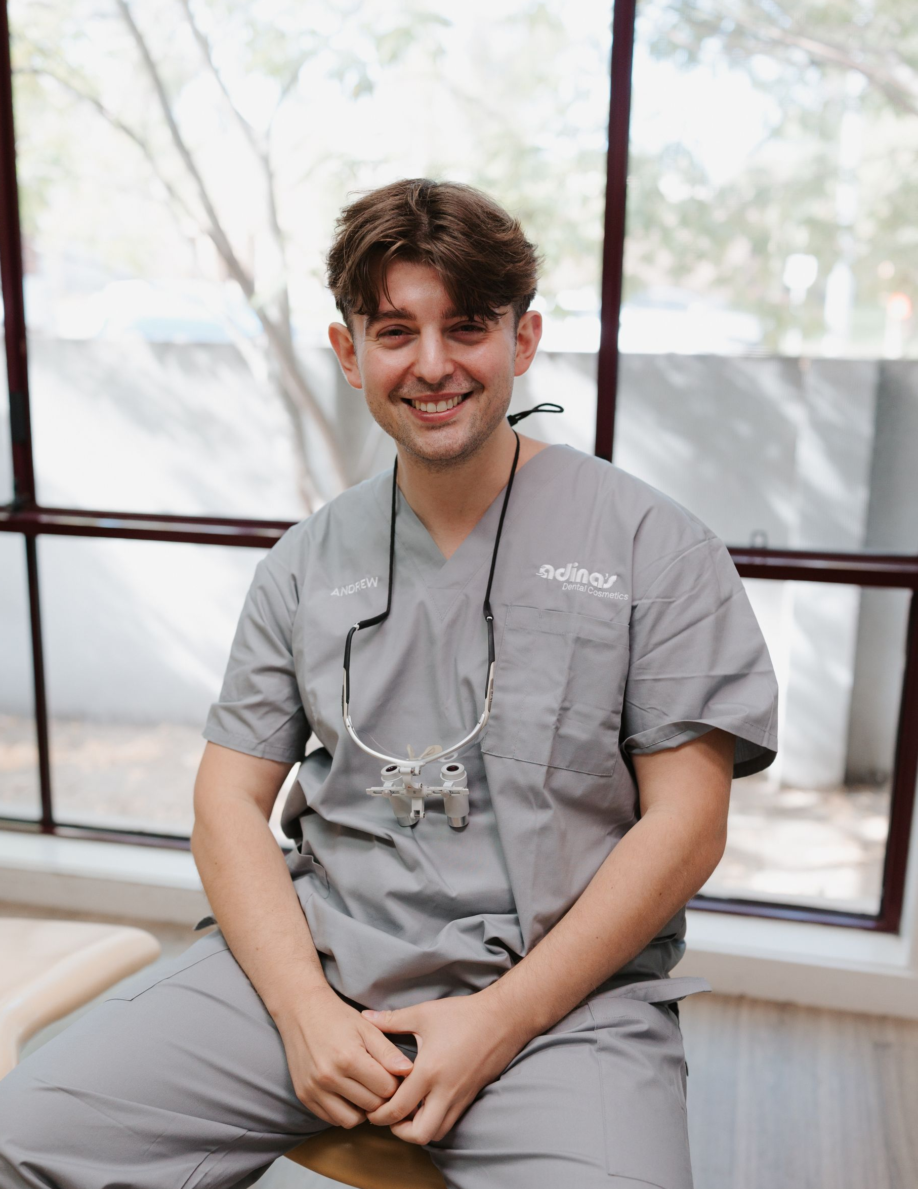 Man wearing grey scrubs, smiling in front of a window.